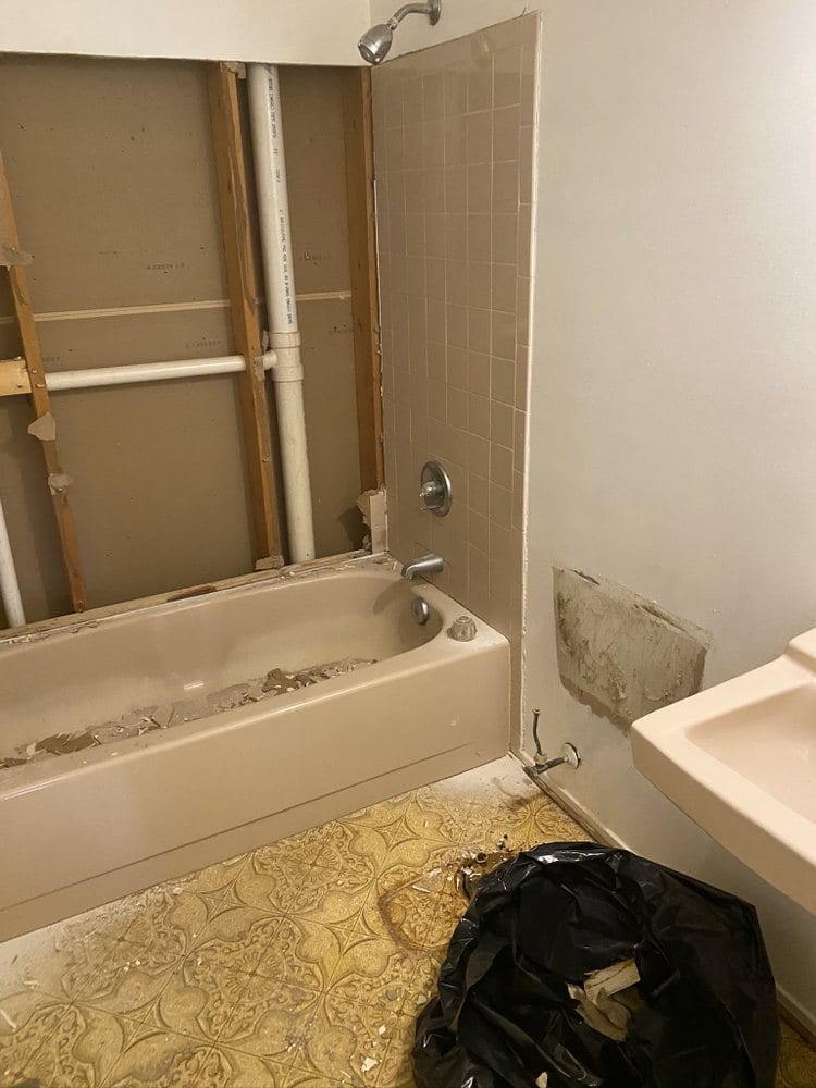 Bathroom mid-demolition with the tub surround removed, exposing wall studs, supply pipes and old vinyl flooring.