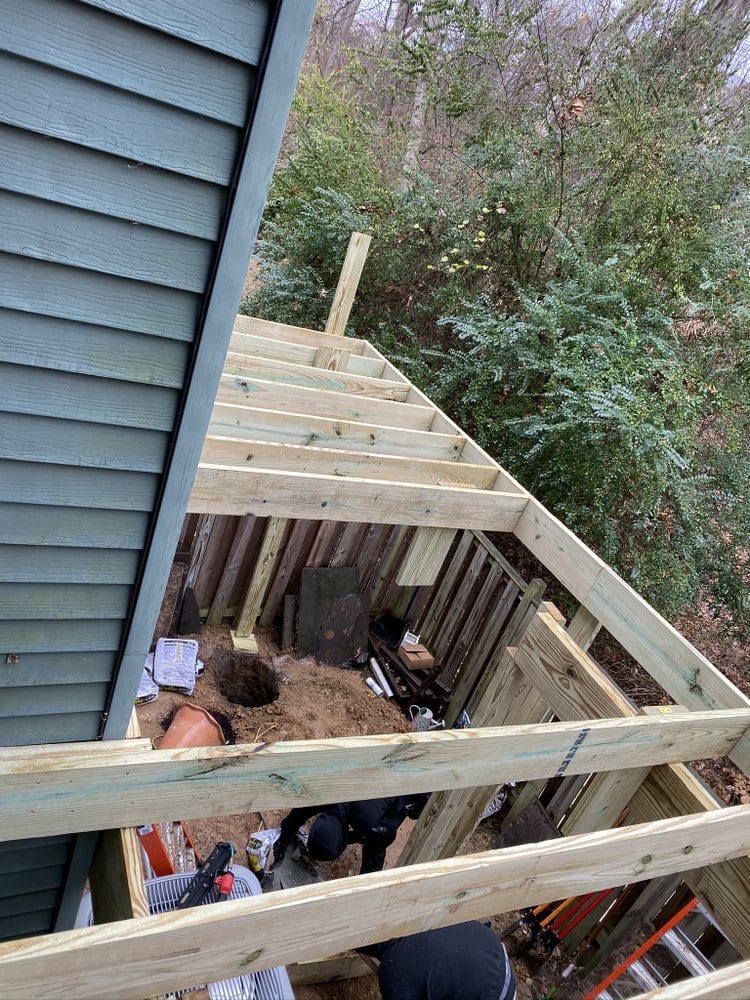 Crew working below new deck framing attached to blue lap siding on a hillside lot.