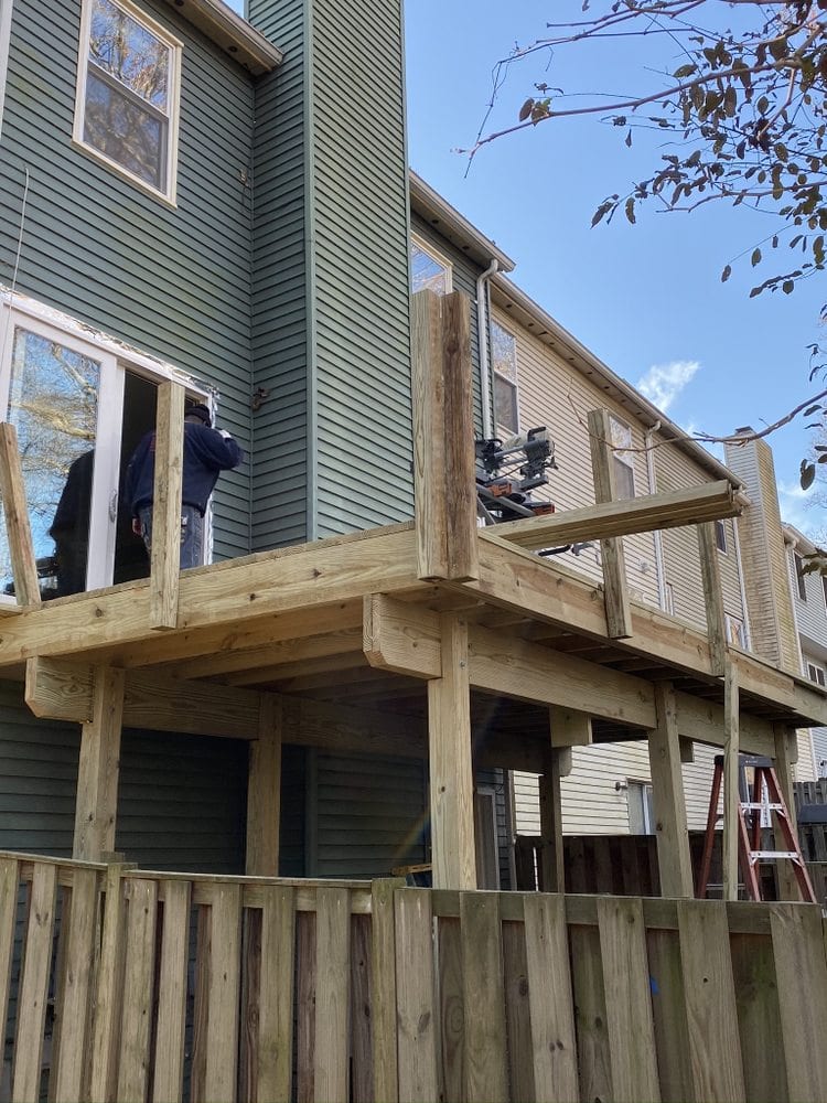 Partly rebuilt deck and railing on a green-sided house, with new lumber replacing old sections.