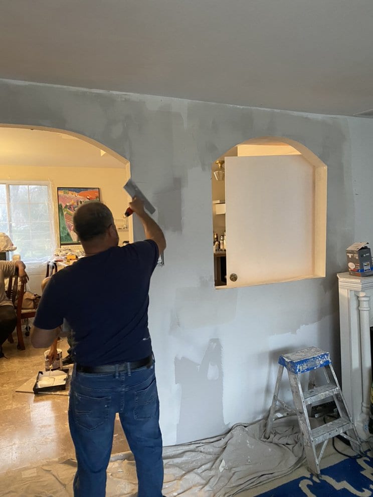 A worker running a wide taping knife over a newly boarded interior wall, applying skim coat beside an arched opening.