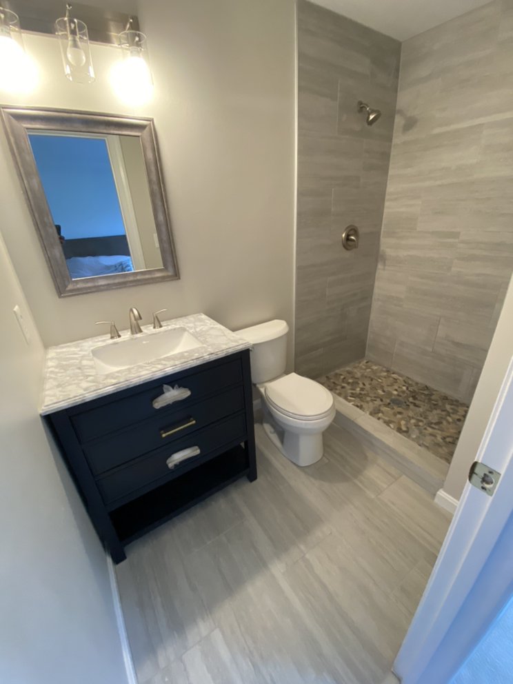 Finished bathroom with a dark navy vanity, framed mirror, toilet and a tiled walk-in shower with a pebble floor.