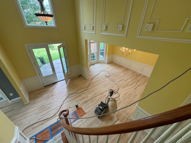 Worker operating a floor sander across an open room with stairs and large windows.