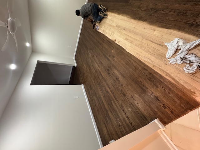 Dark hardwood floor during refinishing with an unfinished section and tools visible.