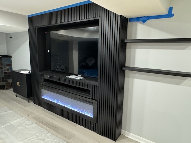 Close view of a black fluted fireplace and media surround with cabinetry and shelf lighting.